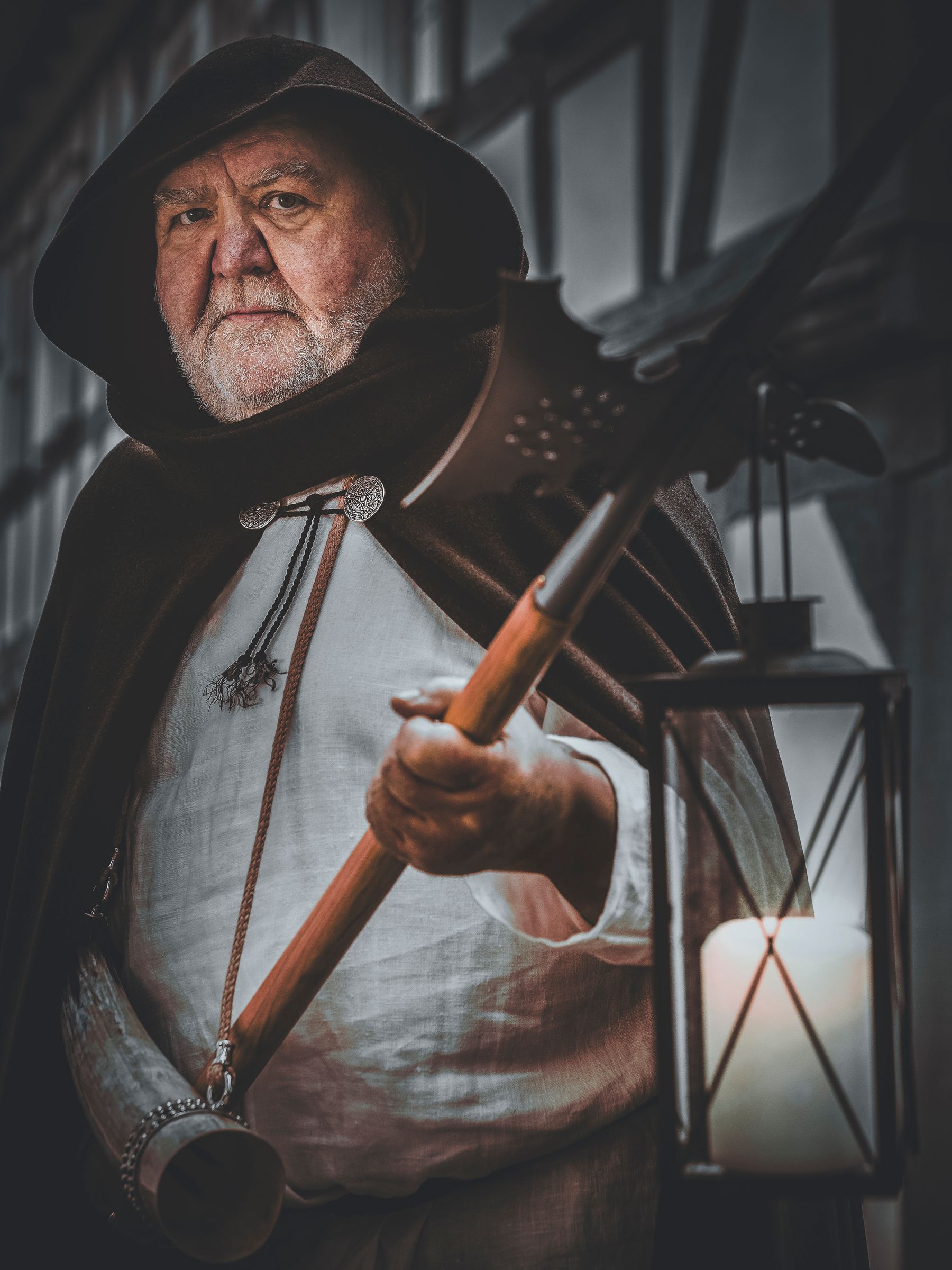 Ein älterer Mann in einem Kapuzenmantel hält eine große Stangenwaffe und eine Laterne mit einer brennenden Kerze. Er steht mit ernster Miene im Freien und trägt eine mittelalterliche Kleidung.